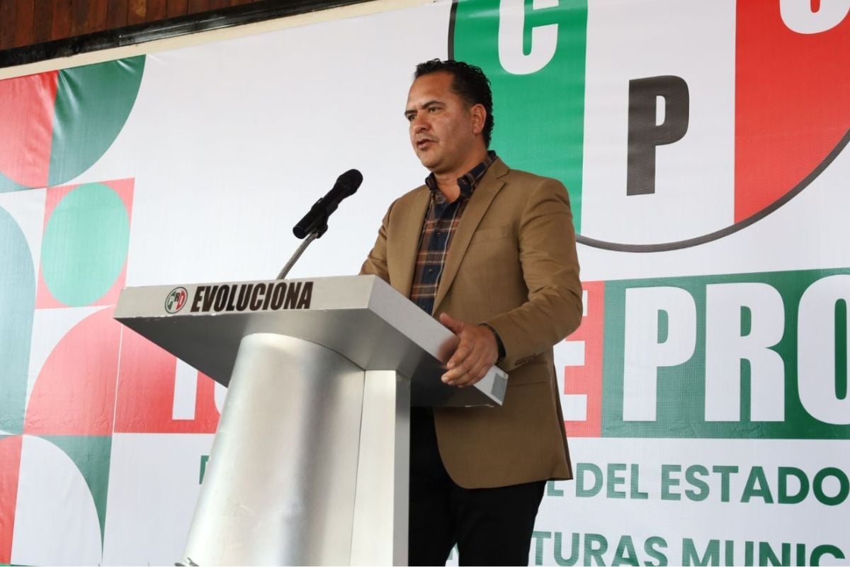 Man in brown blazer speaking at podium with microphone during political event with Mexican flag backdrop