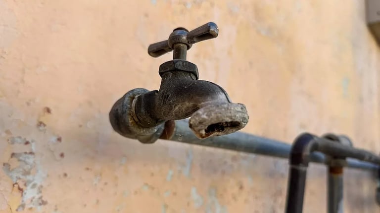An old, weathered metal outdoor water tap mounted on a worn beige wall with visible corrosion and patina