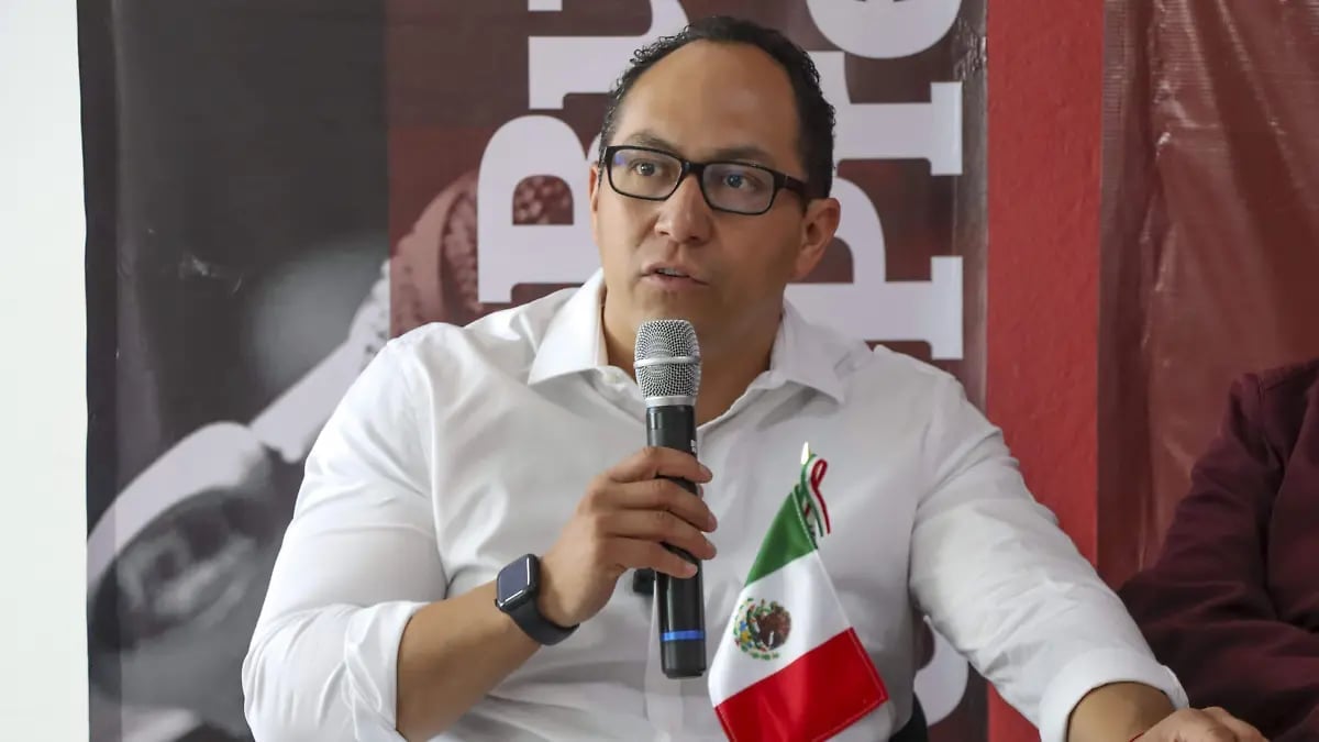Man with glasses in white shirt speaking into microphone with Mexican flag pin visible