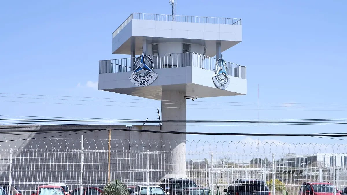 Elevated white box structure with clock faces on a tall pole above a chain-link fence and parking lot