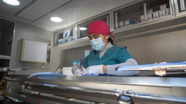 Medical professional in burgundy cap and blue scrubs wearing gloves and mask, working at laboratory equipment in clinical setting