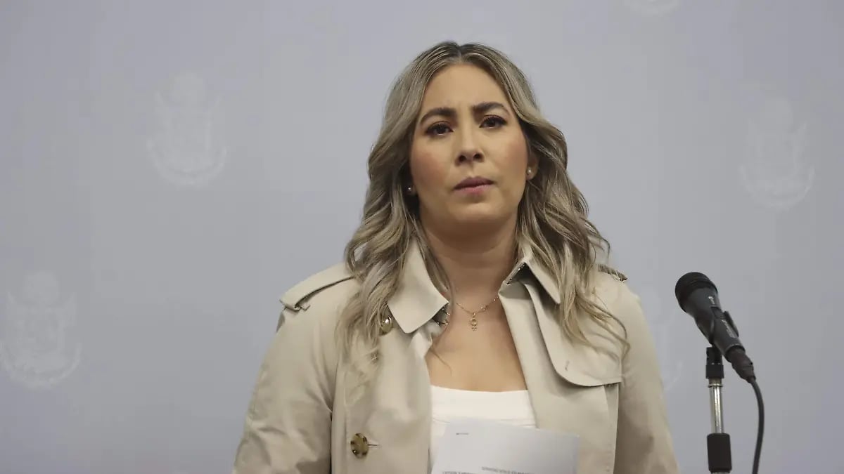 Woman with long blonde hair wearing a beige blazer standing at a microphone against a plain background