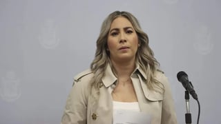 Woman with long blonde hair wearing a beige blazer standing at a microphone against a plain background