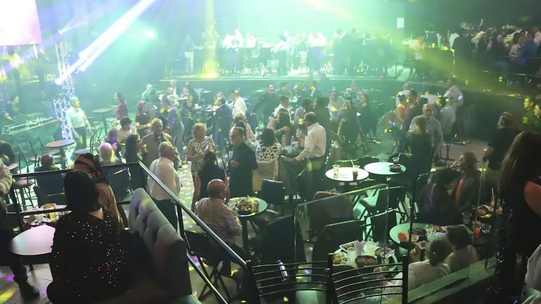Crowded indoor event venue with large group of people dancing and socializing under green stage lighting